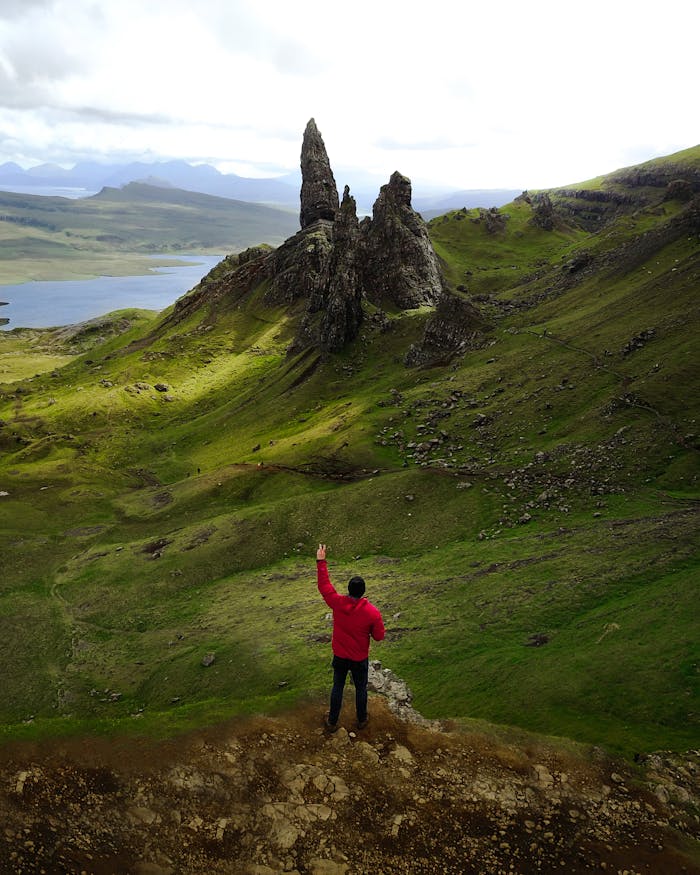 A lone hiker in a red jacket stands before the iconic Old Man of Storr on the Isle of Skye, Scotland.